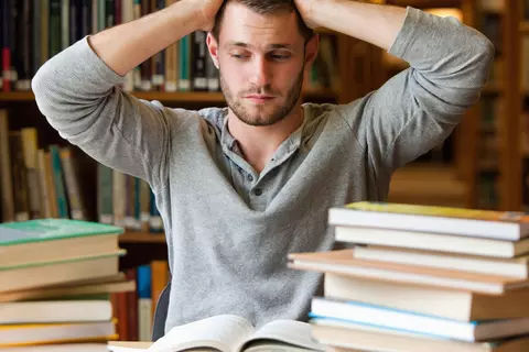 Student am Schreibtisch voller Bücher hält sich die Hände an den Kopf