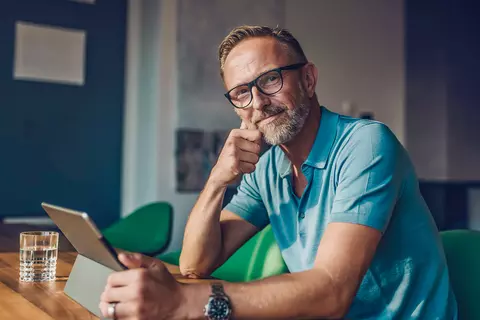 Ein Mann sitzt am Tisch und hält ein Tablet, die andere Hand ruht am Kinn. 