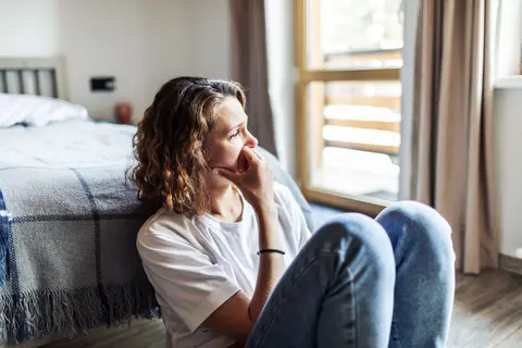 Eine junge nachdenkliche Frau, die allein auf dem Boden vor einem Bett sitzt