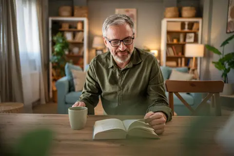 Ein reifer Mann genießt einen ruhigen Moment beim Lesen eines Buches, während er an einem Tisch sitzt und eine Tasse Kaffee vor sich stehen hat.