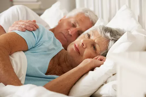 Seniorin liegt wach neben schlafendem Partner im Bett