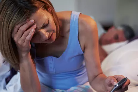 Erschöpfte Frau sitzt im Bett mit Handy in der Hand